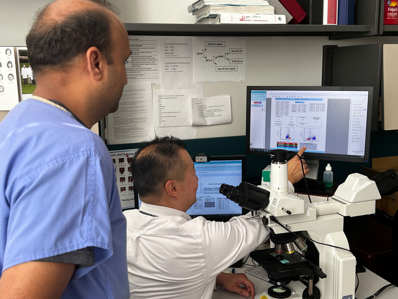 Two people in a lab are examining flow cytometry data on a computer monitor, with a microscope and documents visible on the desk.